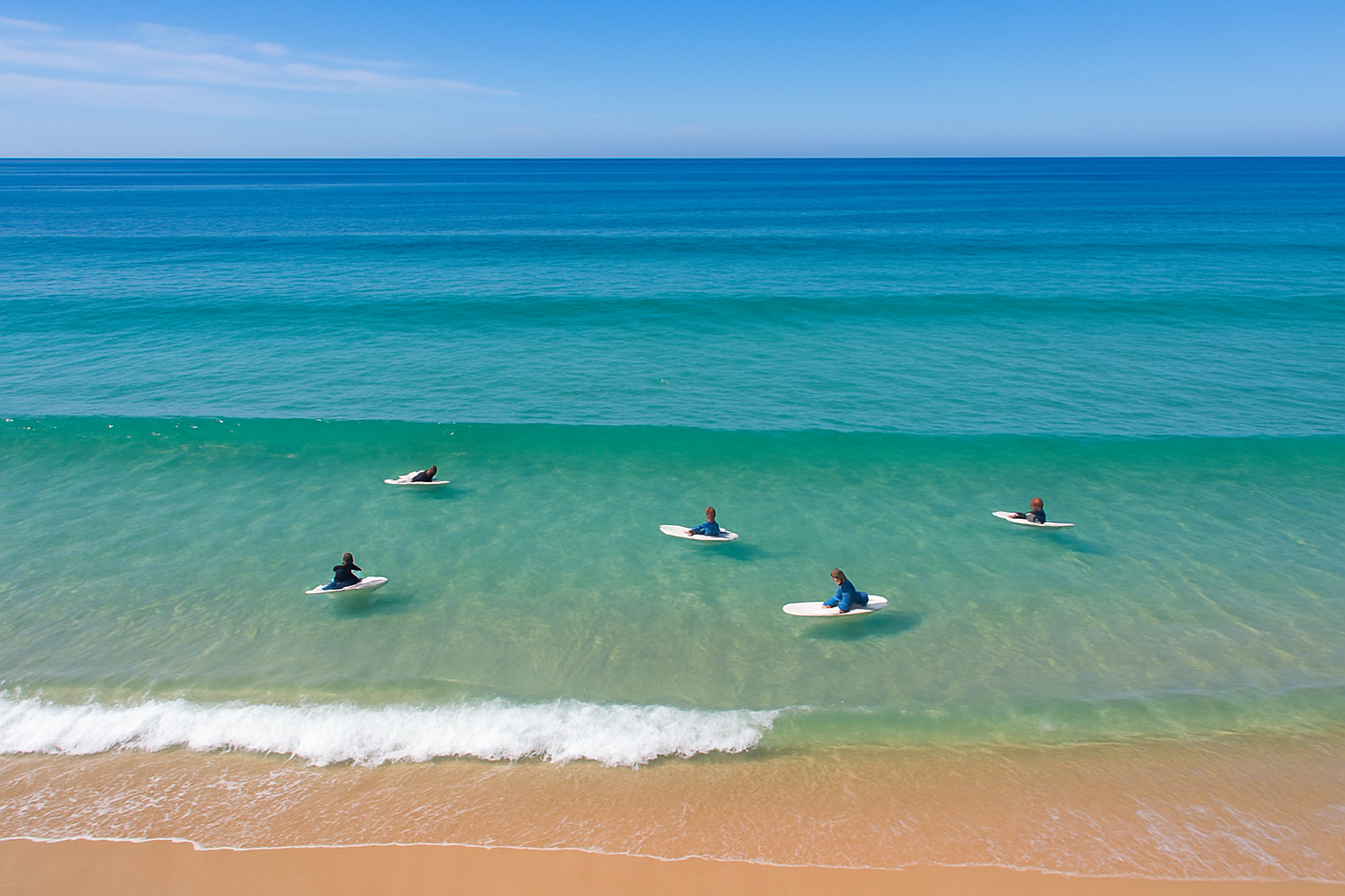 Aerial view of beginner surfers on foam boards paddling in calm shallow waves at a beginner-friendly surf beach