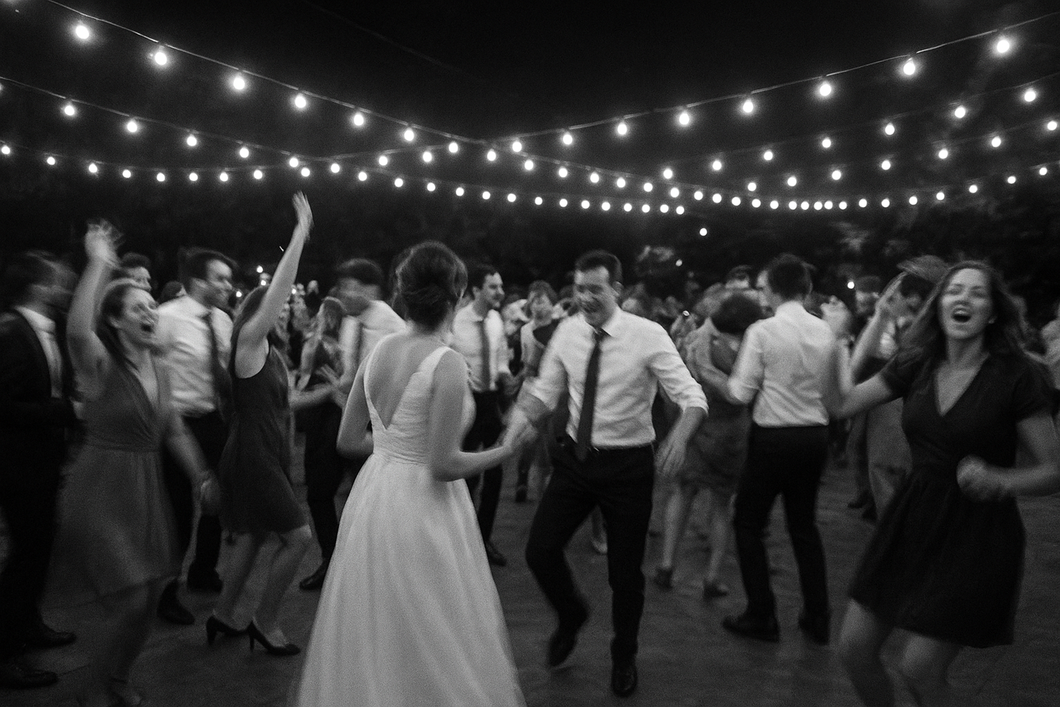 Black and white wedding photo of guests dancing at a reception under string lights